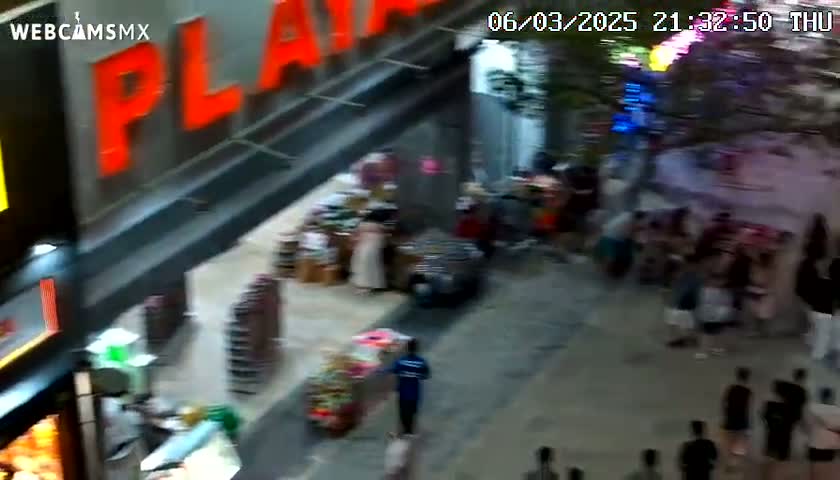 A high-angle, nighttime view of a bustling street scene outside a shopping mall, with people walking around and shopping.