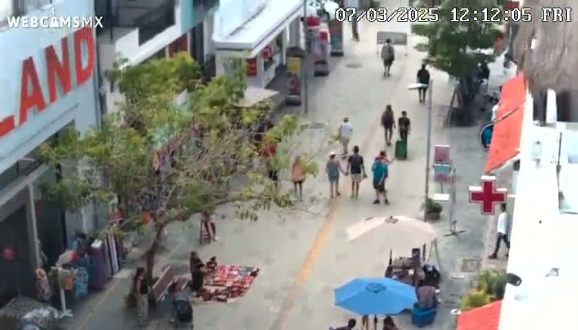 A paved pedestrian street lined with shops and trees, with several people walking around on a sunny day.