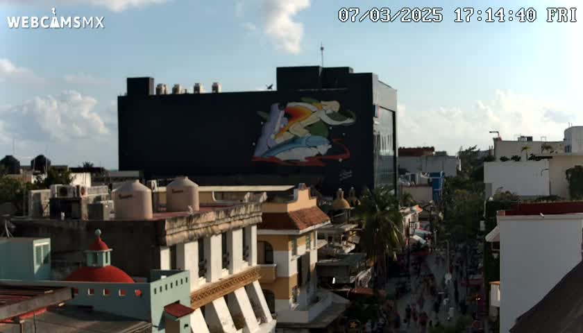A partly cloudy daytime view of a city street scene, featuring a large building with a mural and surrounding low-rise buildings.