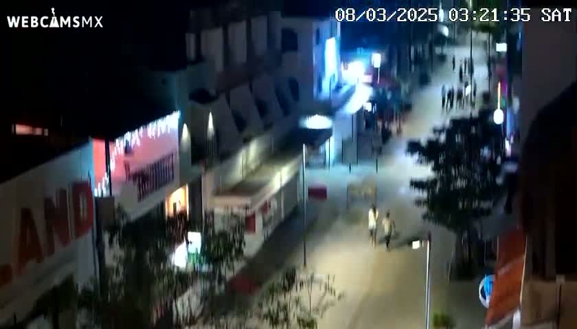 An aerial nighttime view of a street scene shows several people walking past brightly lit buildings and shops.