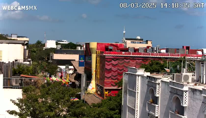 A partly sunny outdoor scene shows a cityscape with several buildings, including a multi-story building with red and yellow chevron patterns, and lush green trees and vegetation scattered throughout.
