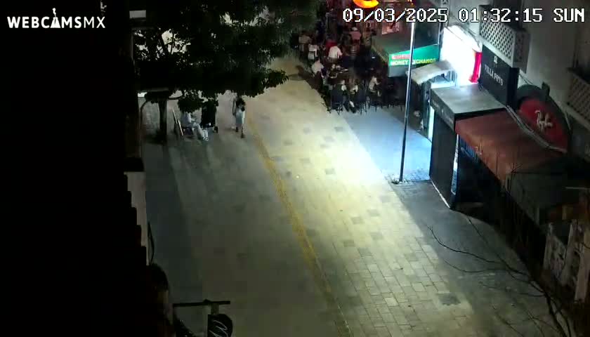 An empty nighttime street, illuminated by artificial light, shows a few pedestrians and some outdoor seating areas of businesses.