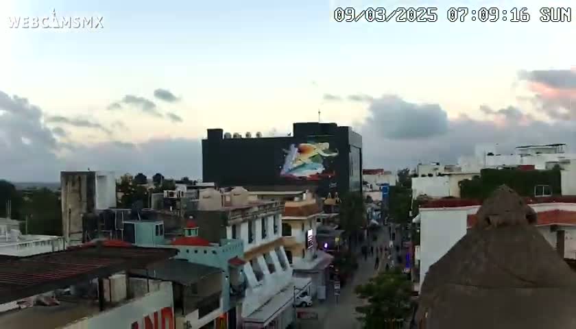 An aerial view of a city at sunrise under a partly cloudy sky shows buildings with a large mural depicting a person on a building's side.