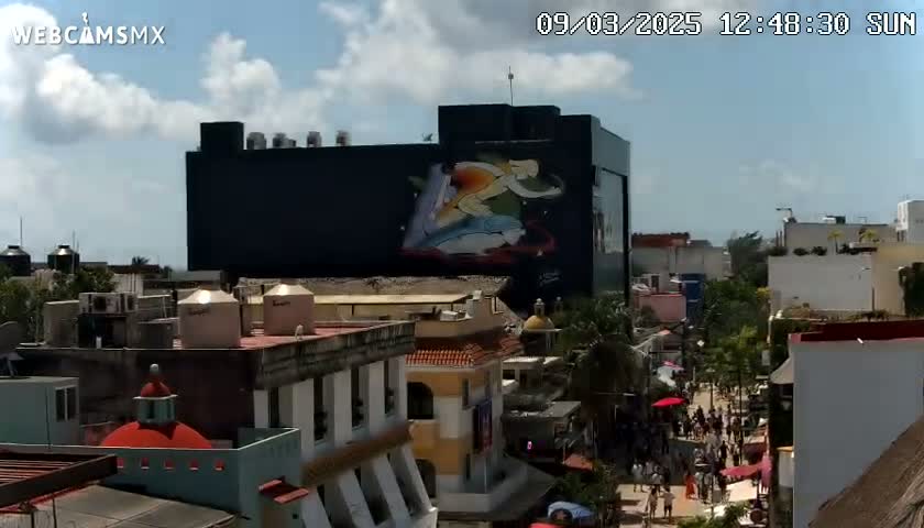 A partly cloudy sunny day reveals a cityscape with a large building displaying a mural, and a busy street scene below.