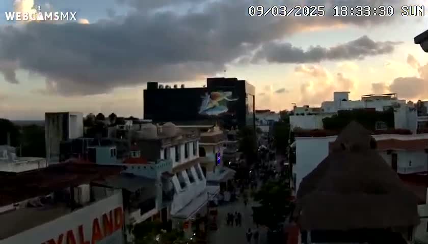 A sunset casts a warm glow over a city street scene with numerous buildings, some with distinctive architectural features, under a partly cloudy sky.