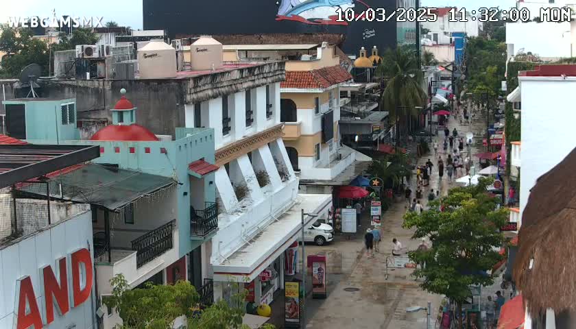 A street scene shows pedestrians walking on a wet street lined with multi-story buildings, some with balconies and awnings.