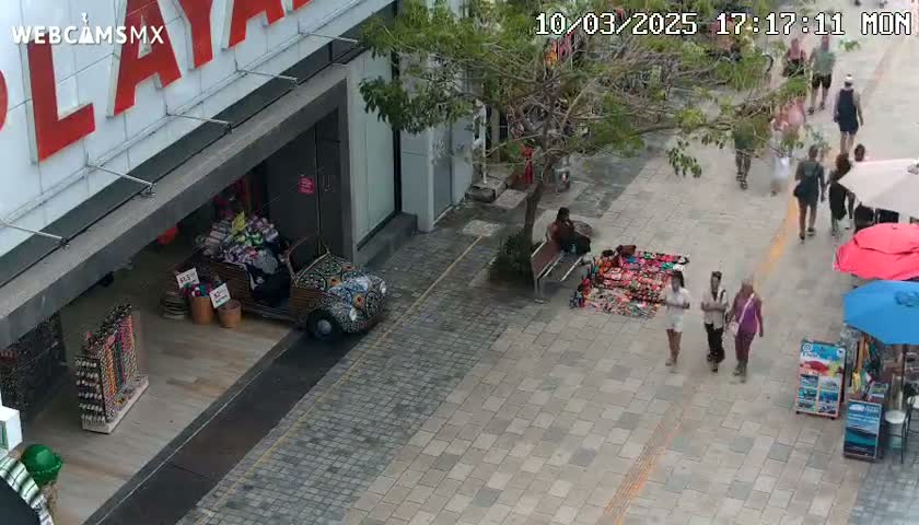 An aerial view of a paved pedestrian walkway shows several people walking past street vendors and a decorated Volkswagen Beetle parked near a store.
