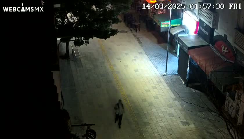 A person walks down a mostly empty, brightly lit street at night.
