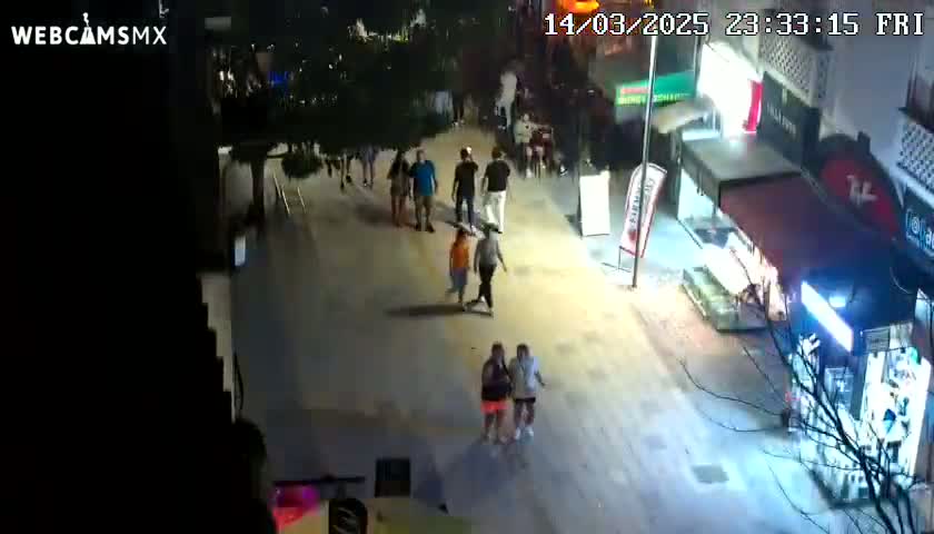 A high-angle nighttime view shows a sparsely populated street with several people walking near shops and businesses under partly cloudy skies.