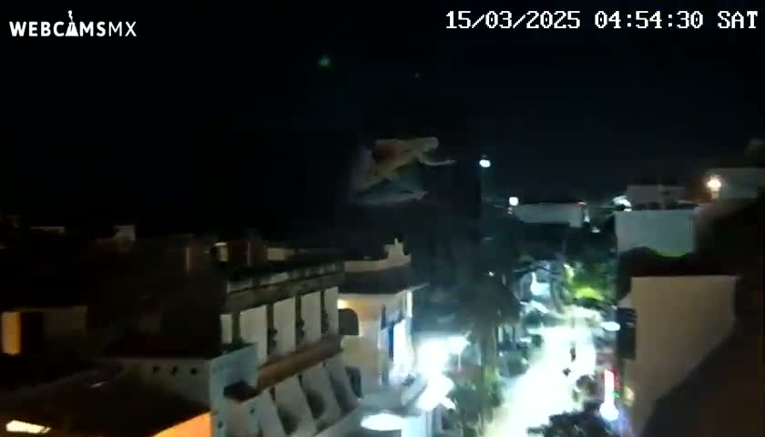 A nighttime aerial view shows a person falling from above a street lined with buildings and palm trees under a clear sky.