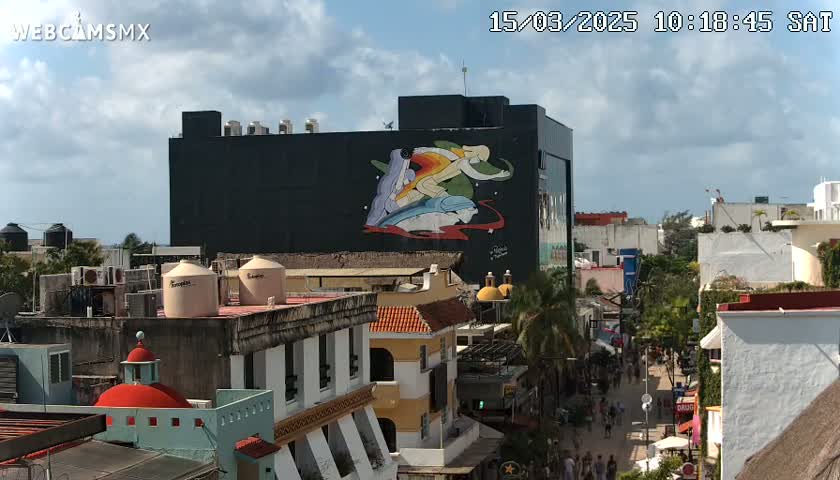 A partly cloudy day reveals a cityscape with colorful buildings, a large mural depicting stylized figures and dolphins on a building, and a street scene below.
