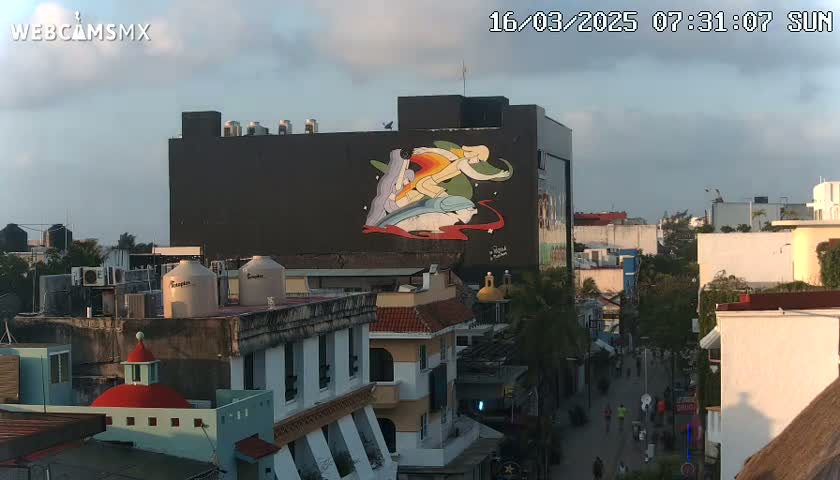A large building with a colorful mural depicting figures in motion is visible from an elevated viewpoint, overlooking a city street with pedestrians and other buildings under a partly cloudy sky.