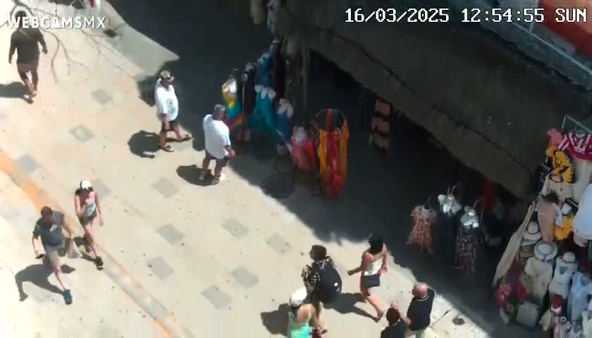 A sunny day shows several people walking on a paved walkway beside various clothing displays and vendors.
