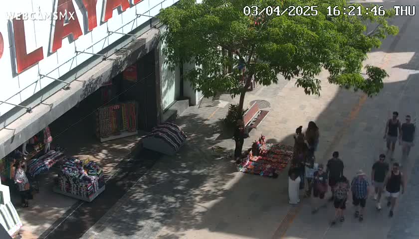 A sunny outdoor street scene shows several people walking and shopping near a building with a large sign and vendors displaying merchandise on blankets on the sidewalk.