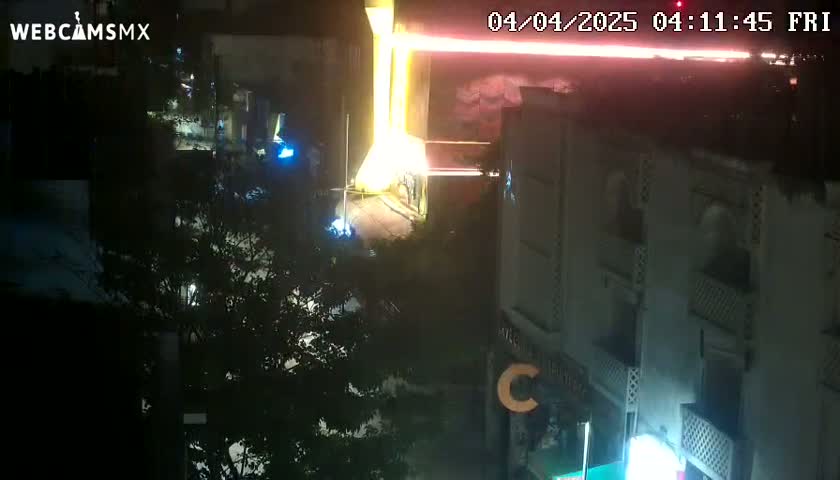 A nighttime aerial view shows a street scene with brightly lit buildings and trees, illuminated by what appears to be fireworks in the distance.