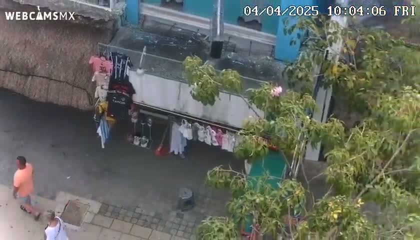 An overhead view shows a street scene with a man and a woman walking past a small shop displaying clothing, partially obscured by a leafy tree on a sunny day.