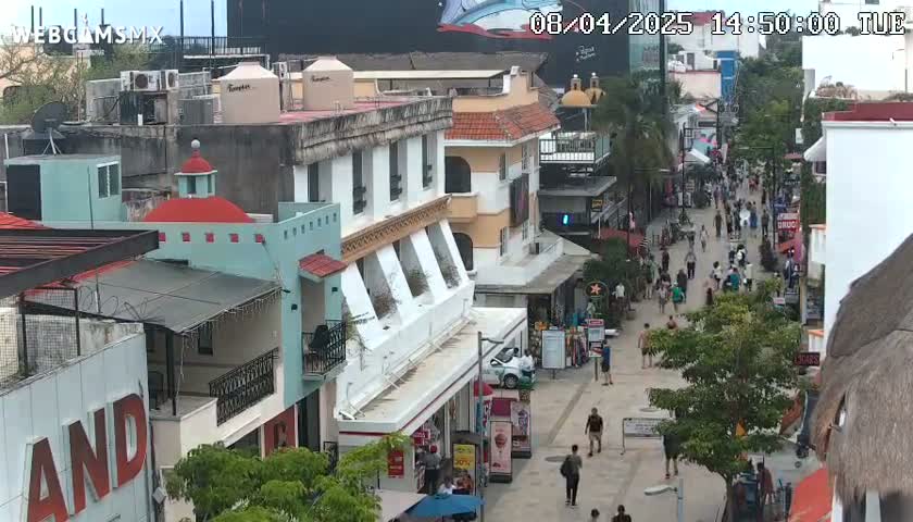 A pedestrian street scene in a town with multi-story buildings, many people walking, and a partly sunny day.