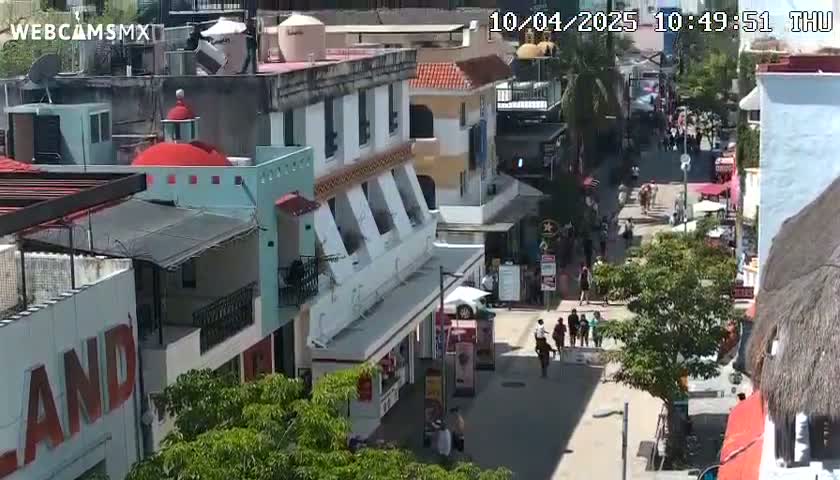 A sunny, daytime view of a pedestrian street lined with colorful buildings, featuring several people walking and shops.