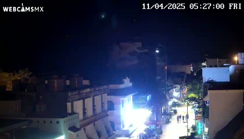 A nighttime, overhead view of a street scene in a town or city with buildings lit up, a few people walking on the street, and a clear sky.