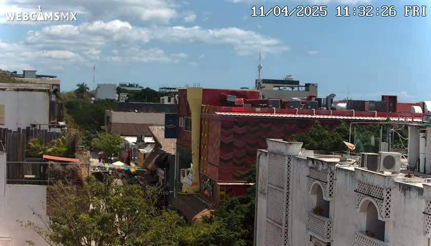 A partly cloudy day reveals a cityscape of multi-story buildings, some with vibrant facades, nestled amongst lush green trees.