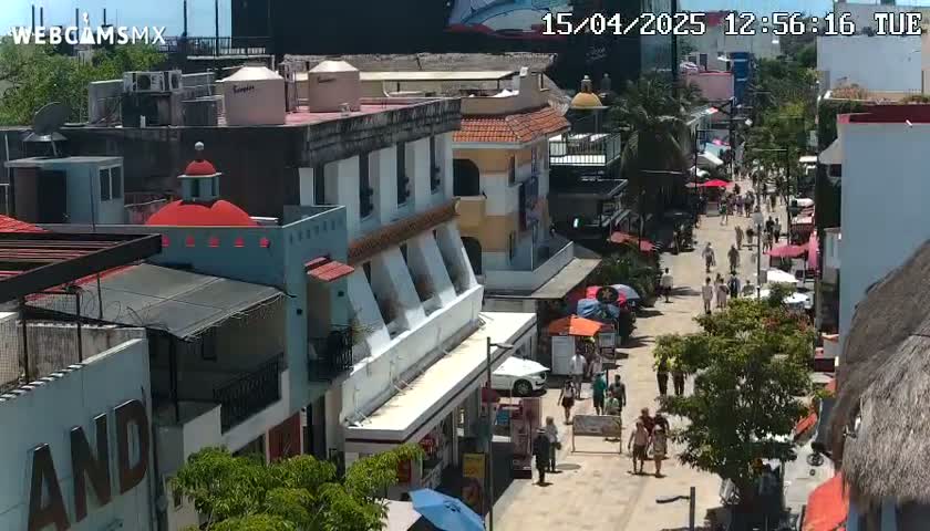 A sunny street scene shows a pedestrian walkway lined with multi-story buildings and numerous people walking.