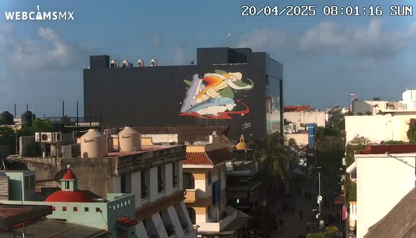 A city street scene under a partly cloudy sunny sky features a large building with a mural depicting stylized figures on its side, surrounded by other buildings of varying heights and architectural styles.