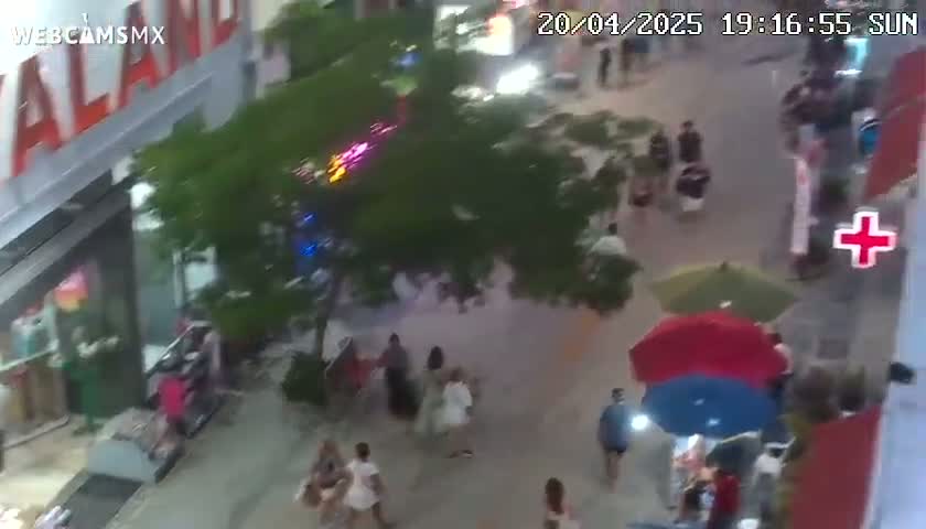 An outdoor pedestrian area at night is shown with people walking past shops and restaurants under a few umbrellas.