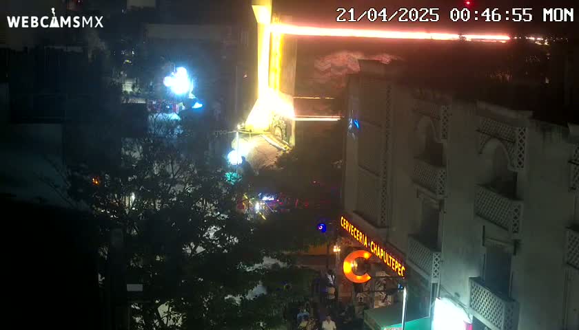 A nighttime aerial view of a city street shows brightly lit buildings and trees, with some people visible near a restaurant.