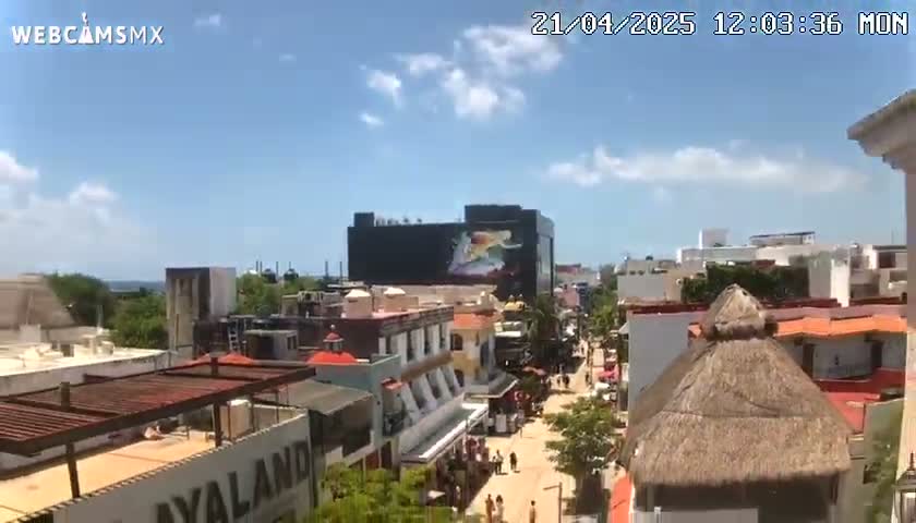 A partly sunny day reveals a view of a tropical city street with colorful buildings, pedestrians, and a large mural on a building in the background.