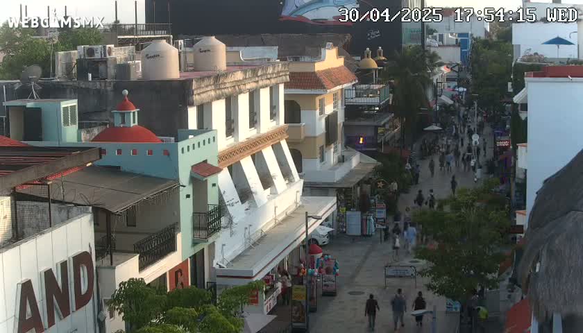 A pedestrian street scene at dusk shows several multi-story buildings, shops, and many people walking along a busy street.