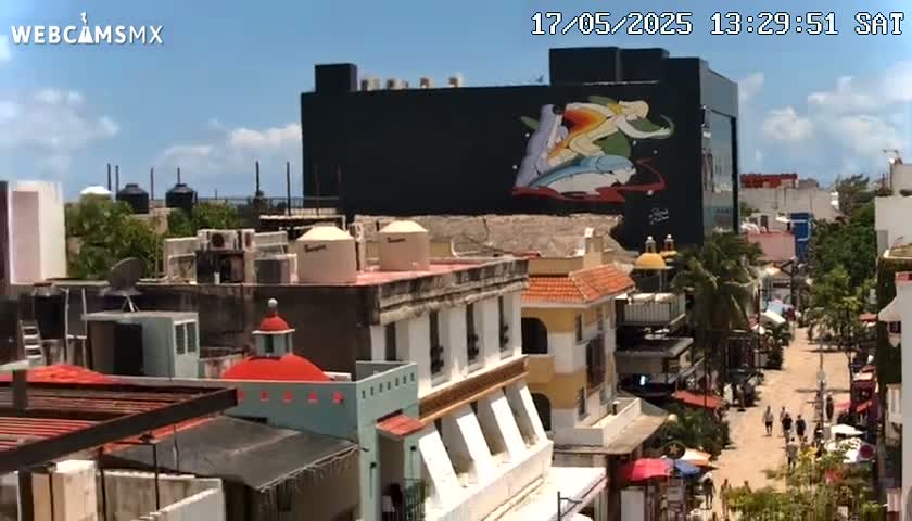 A sunny day reveals a cityscape of low-rise buildings, one prominently displaying a large mural, with pedestrians visible in a street below.
