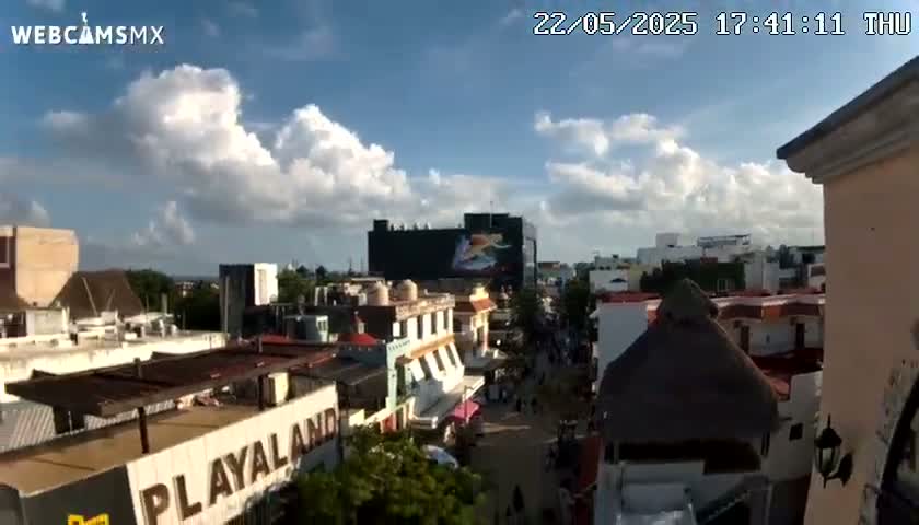 A partly cloudy daytime view of a city street scene, featuring buildings with varied architectural styles, including a large building with a billboard, under a partly cloudy sky.