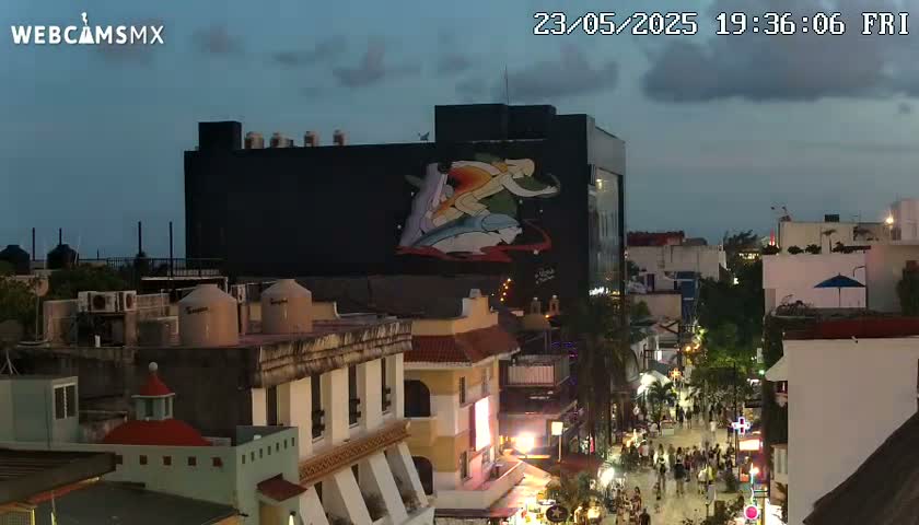 A nighttime, dusk-lit cityscape featuring a large building with a mural depicting figures on a wave, overlooked by other buildings and a street filled with pedestrians.
