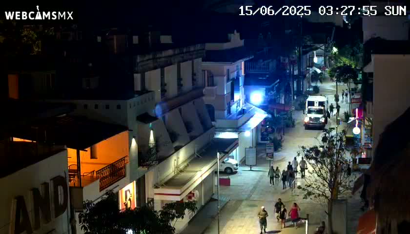 A nighttime, high-angle view shows a street scene in a town with pedestrians and a van, with buildings lit up along both sides of the street.