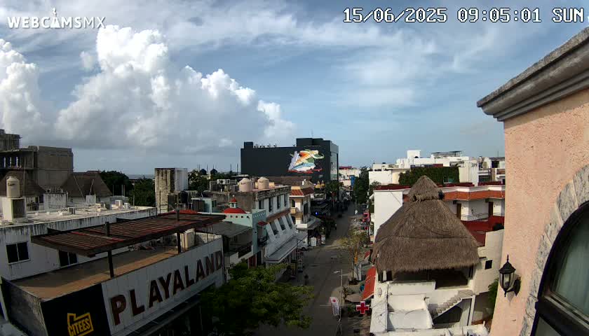 A partly cloudy sunny day overlooks a city street scene with low-rise buildings, a large mural on one building, and a thatched roof structure.