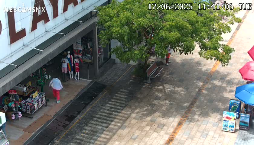 A sunny outdoor scene shows a paved walkway with a person walking past a storefront displaying mannequins, a park bench, and several umbrellas.