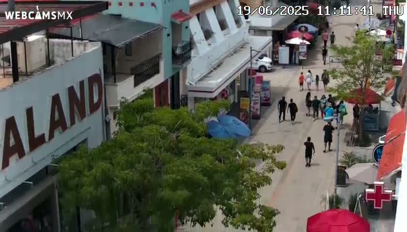 A high-angle view shows a pedestrian street scene with numerous people walking among buildings, under a sunny sky.