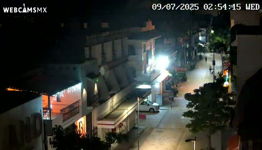 A nighttime, overhead view of a mostly empty street lined with brightly lit buildings.