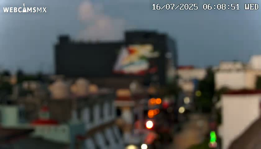 A blurry, dusk image shows a city street with buildings and streetlights, under a mostly cloudy sky.