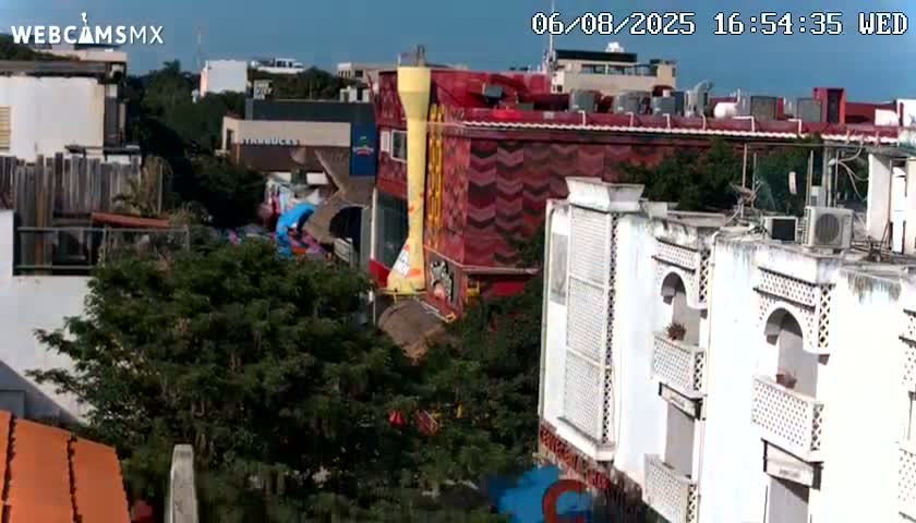 An aerial view shows a street scene with colorful buildings, lush green trees, and a clear blue sky.