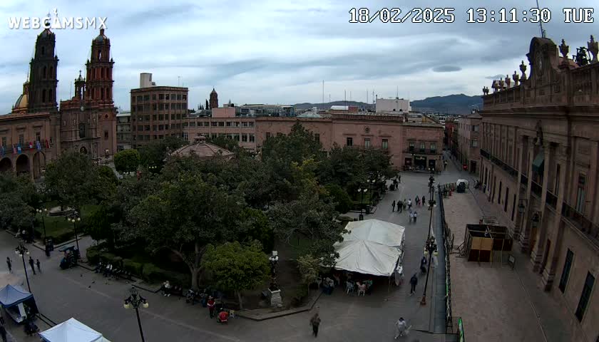 An elevated view of a city square with a cathedral, other buildings, and lush green trees under a cloudy sky.