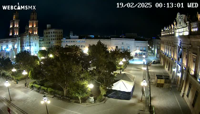 A nighttime aerial view of a city square, featuring a cathedral, other buildings, lush trees, and a covered structure in the center, under clear skies.