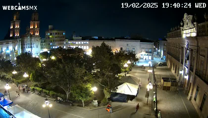 A nighttime, high-angle view of a town square with a cathedral and other buildings illuminated, surrounded by trees and some people.
