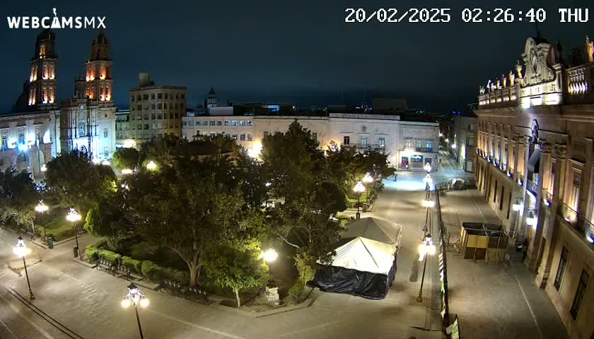 A nighttime, high-angle view of a town square with a cathedral, buildings, trees, and a covered tent, illuminated by streetlights.