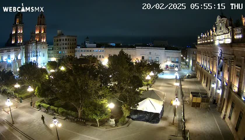 A nighttime aerial view of a plaza with a cathedral and other buildings, illuminated by streetlights and surrounded by trees under a clear sky.