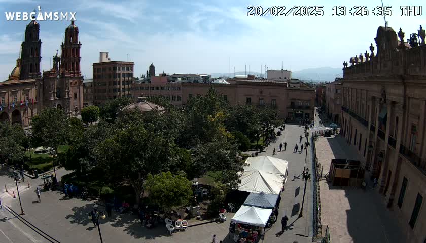 A city square with a park, buildings, and a few people, under a mostly sunny sky.