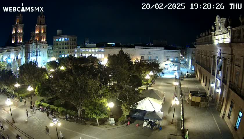 A nighttime aerial view of a city square, illuminated by streetlights, featuring a large cathedral, other buildings, and a park with trees and people.