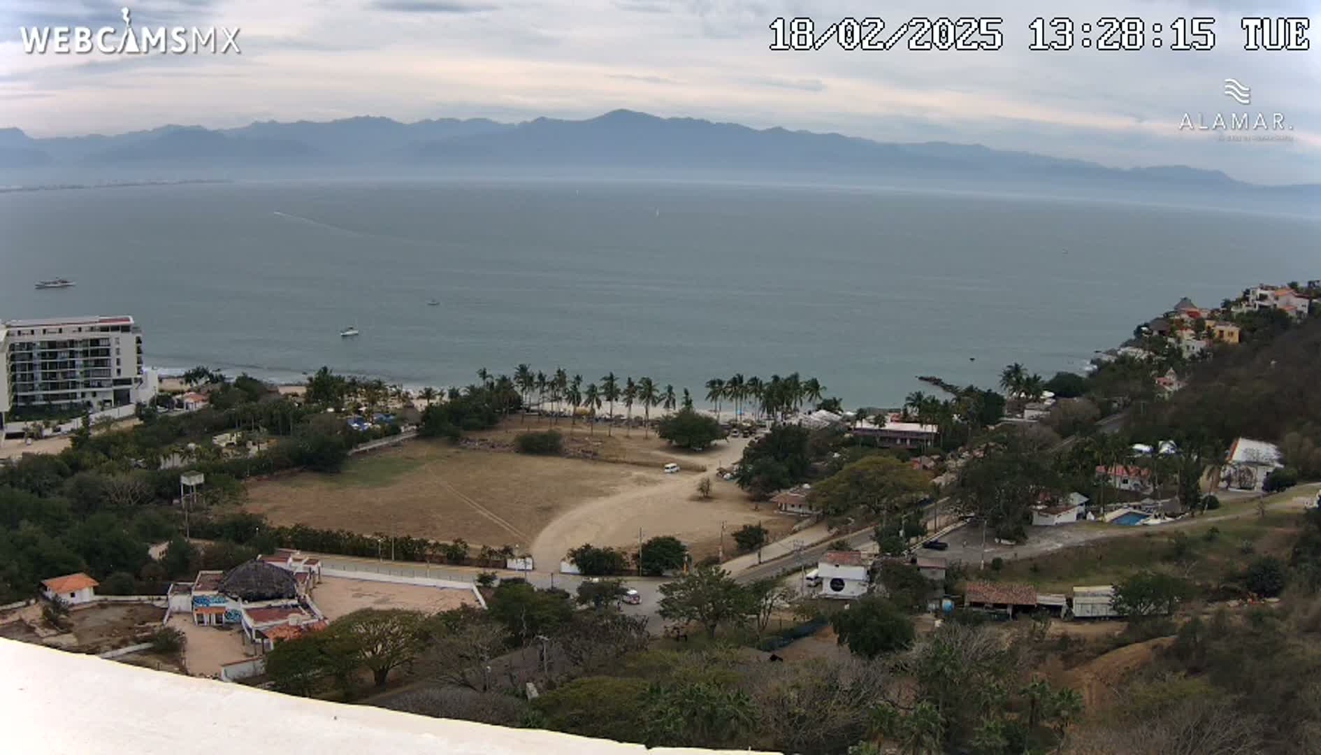 A high-angle view of a coastal town on a cloudy day, showing a beach with palm trees, buildings, and a large body of water with mountains in the distance.