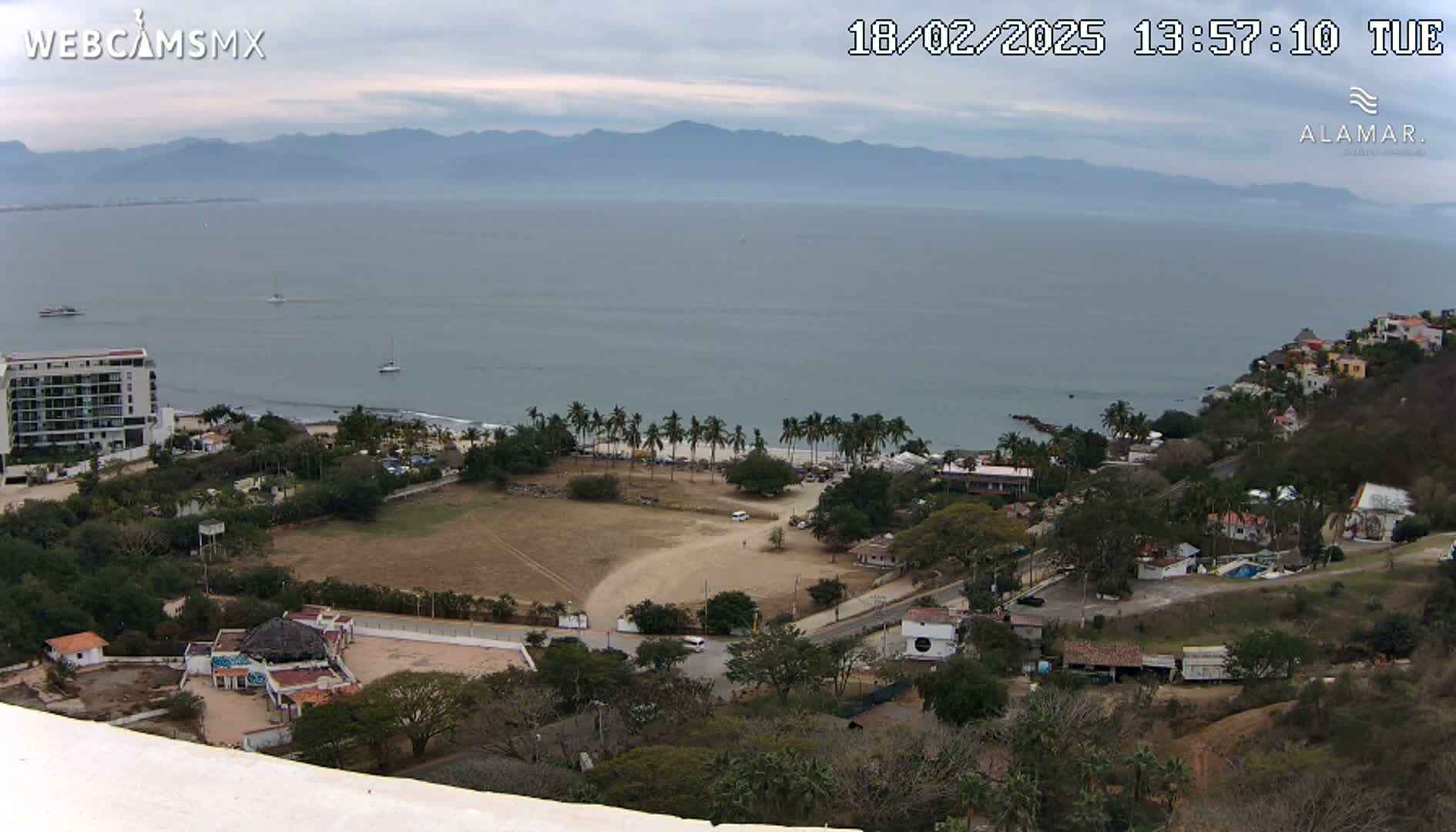 A high-angle view shows a coastal scene under overcast skies, featuring a sandy beach with palm trees, a few buildings, and a calm ocean with distant mountains.