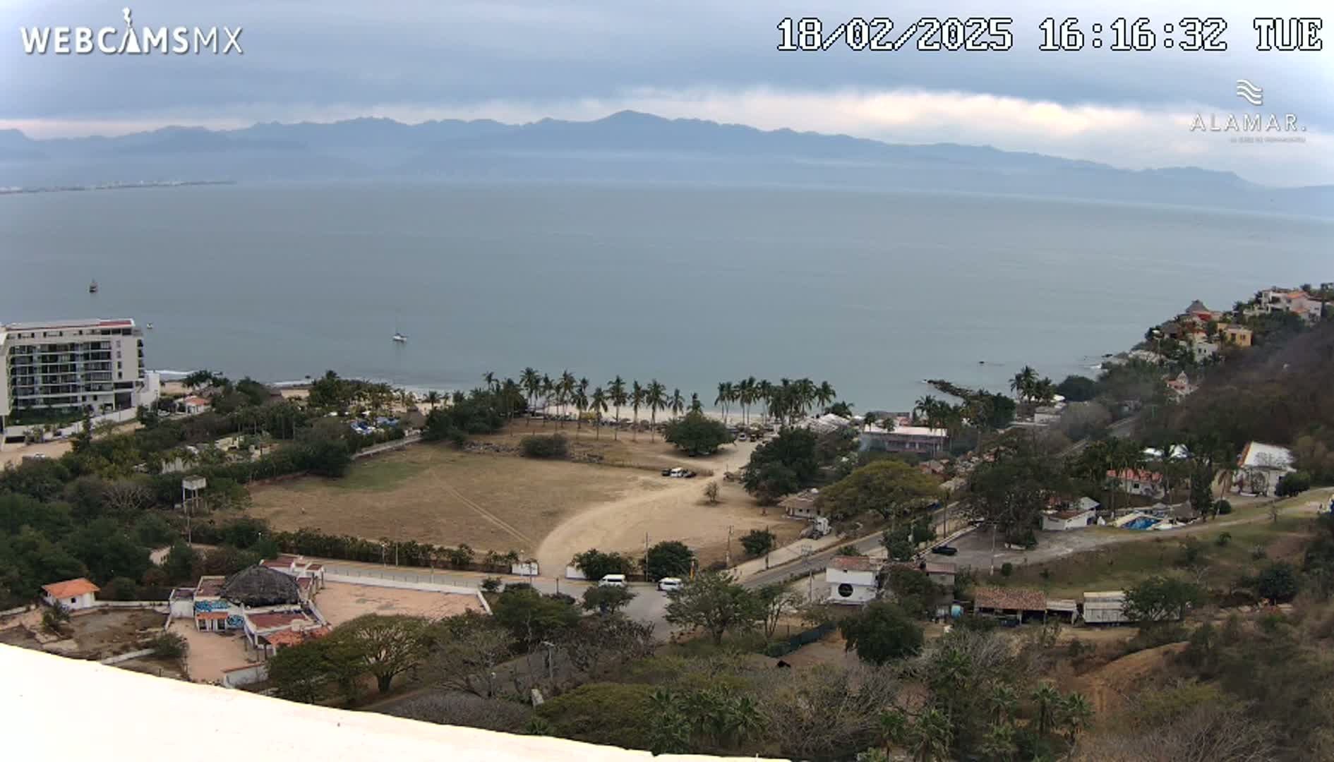 An elevated view of a coastal town on an overcast day, showing a calm ocean, a sandy beach lined with palm trees, buildings, and dry, brown land.